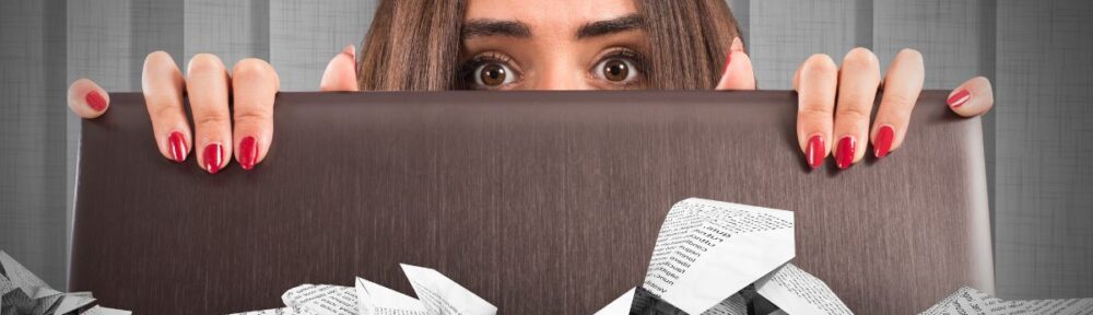 A woman peeking behind a laptop with tons of paper junk